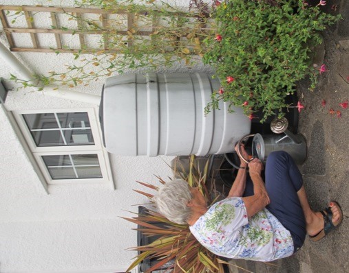 A woman is crouched down filling her watering can from a water but. In the foreground there are plant pots filled with colourful flowers and foliage.